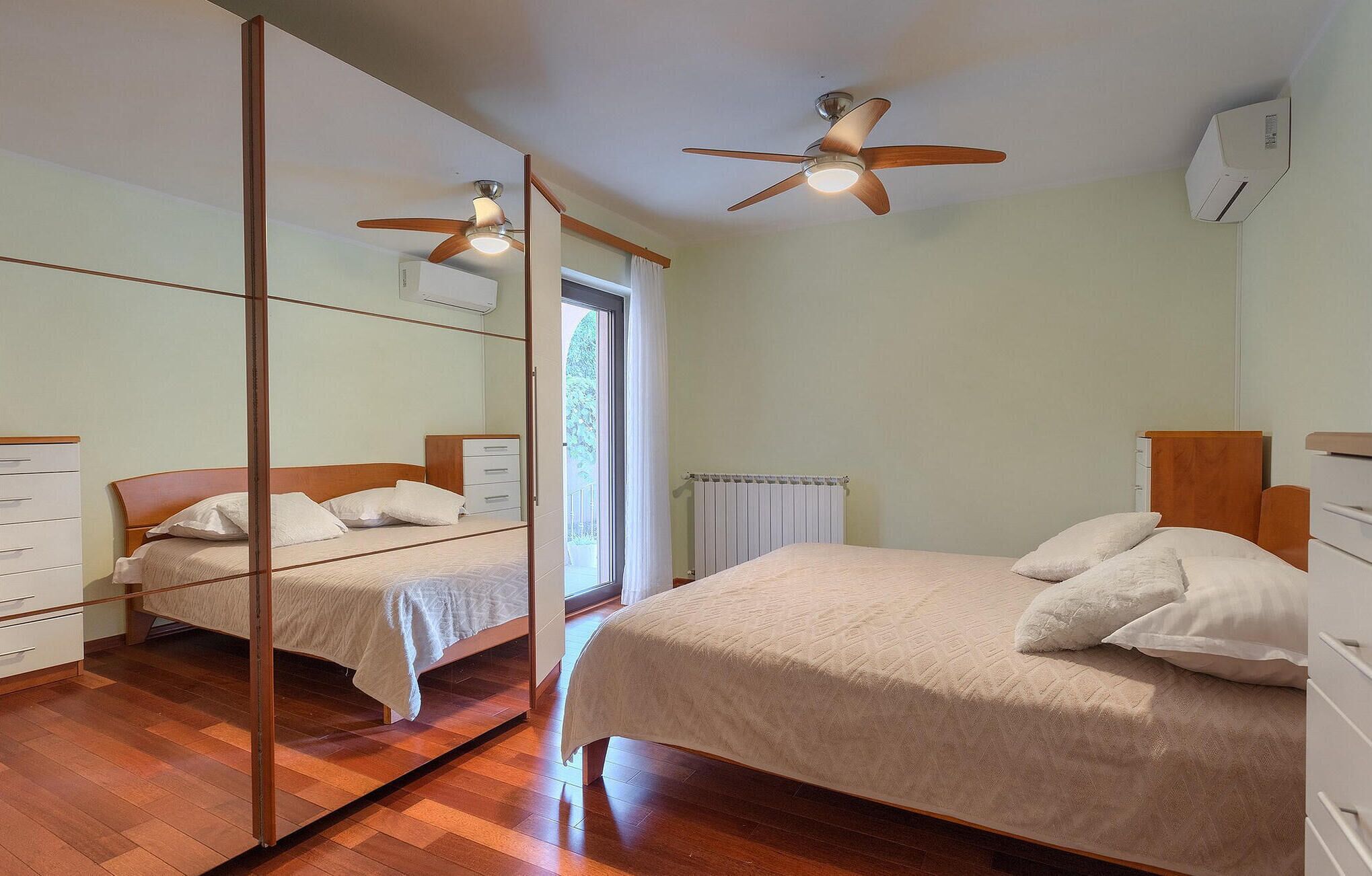 A bedroom with two beds, wooden flooring, a ceiling fan, a radiator, and a window with curtains.