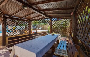 A covered outdoor dining area with a long table and benches, surrounded by wooden lattice walls and a view of a pool and garden. - Awesome home in Pula with sauna (Pula)