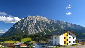 A mountain with a large peak, a yellow and white building with multiple windows, and several smaller houses in the foreground. - Appartement Grimming - Kolb (Tauplitz)