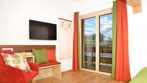 A room with a red chair, a green cushion, a wooden bench, a flat-screen TV, and a sliding glass door with red curtains. - Appartement Grimming - Kolb (Tauplitz)