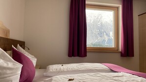 Ein Hotelzimmer mit zwei Betten, einem Holzkopfende, einem Fenster mit Vorhängen und Blick auf die Berge.