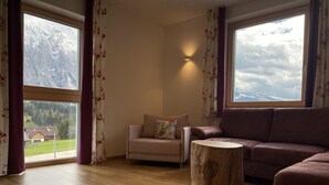 Ein Wohnzimmer mit einer Couch, einem Sessel und einem Holztisch. Große Fenster mit Vorhängen bieten einen Blick auf eine gebirgige Landschaft.