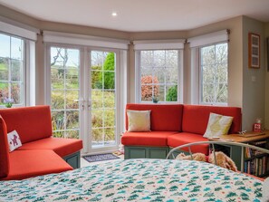 A cozy room with a red sofa, a bed with a patterned bedspread, and large windows offering a view of a garden. - 3 bedroom accommodation in New Galloway (New Galloway)