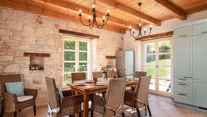 Un coin repas avec des murs en pierre, des poutres en bois au plafond et un lustre. On y trouve des chaises en osier, une table en bois et un réfrigérateur à l’arrière-plan.