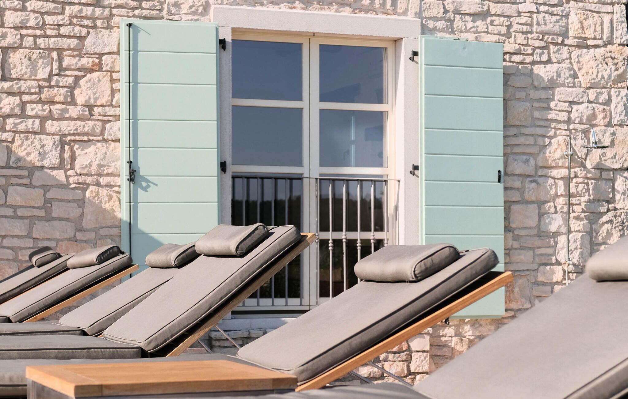 A row of sun loungers with grey cushions against a stone wall, a window with blue shutters, and a metal railing.