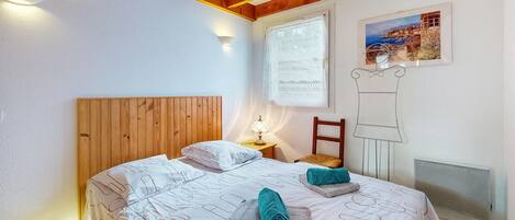 A bedroom with a wooden headboard, a bed with patterned bedding, a nightstand with a lamp, a chair, a radiator, and a framed picture on the wall.
