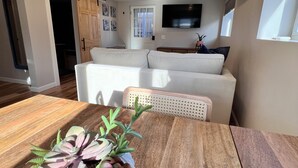 A modern living area with a wooden dining table, a white sofa, and a flat-screen TV mounted on the wall. - Beach Bunker Minute Walk To Beach and Pier at Old Orchard Family-Friendly Cozy (Old Orchard Beach)