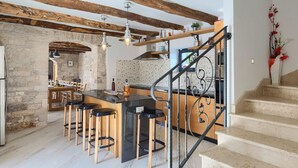 Une cuisine avec des poutres apparentes, un îlot central doté de tabourets de bar et un escalier avec une rampe en fer forgé.