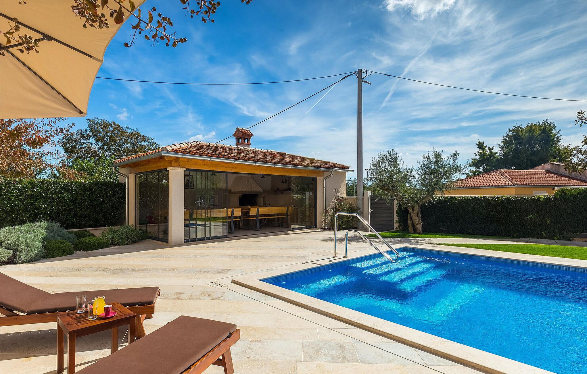 Uma área à beira da piscina com um espaço de estar coberto, a própria piscina e uma casa ao fundo.