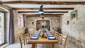 Un espace repas avec une longue table en bois dressée pour le repas, des murs en pierre et des poutres apparentes au plafond.