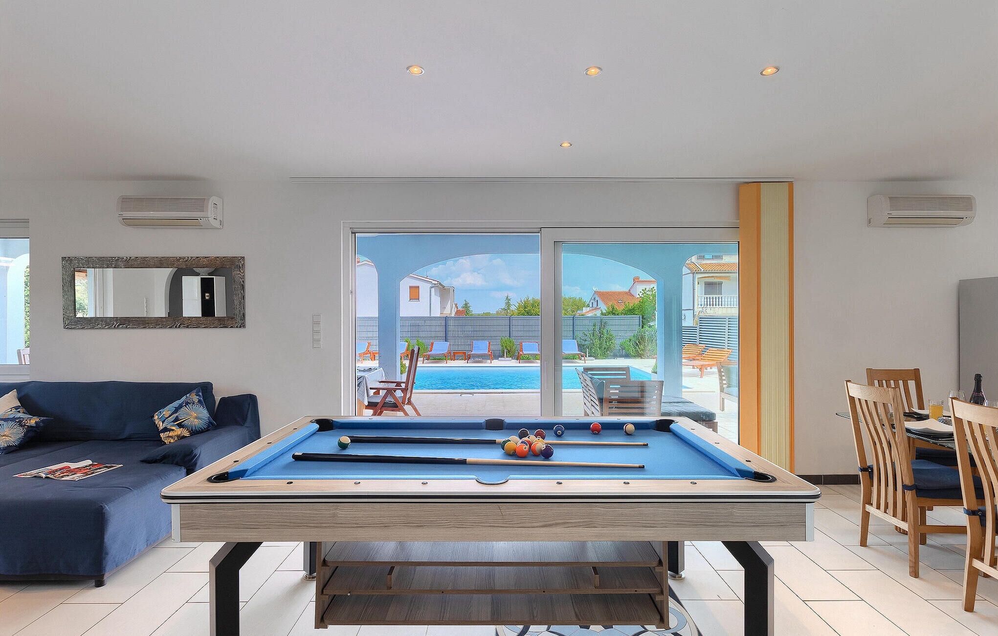 A pool table in a living room with a view of a swimming pool and houses.
