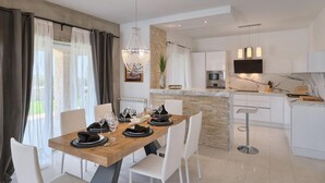 Ein moderner Essbereich mit einem Holztisch, gedeckt für vier Personen, weißen Stühlen, einer Chandelierleuchtre und einer dahinterliegenden Küche mit einer Wand aus Stein.