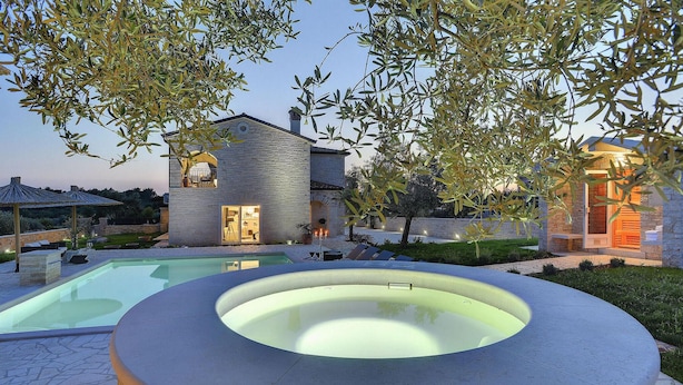 Ein Swimmingpool mit Whirlpool im Vordergrund, ein Steinhaus mit Balkon und Olivenbäume im Hintergrund.