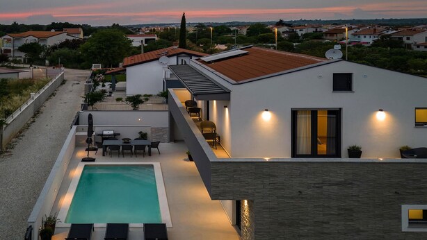 A modern house with a swimming pool, outdoor seating, and a view of the neighborhood during sunset.