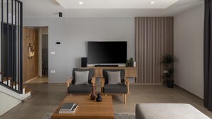 A modern living room with a flat-screen TV, two grey sofas, a wooden coffee table, and a grey armchair.
