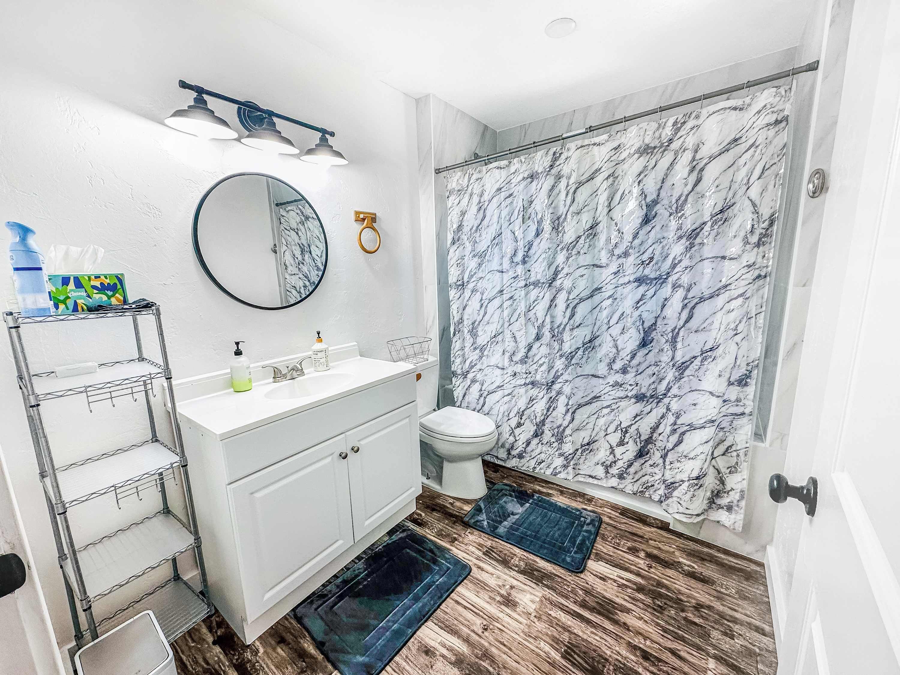 Een badkamer met een witte wastafel, een ronde spiegel, een douche met marmeren wanden en een houten vloer.