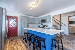 A kitchen with a central island, bar stools, a red door, and a staircase leading to another room.