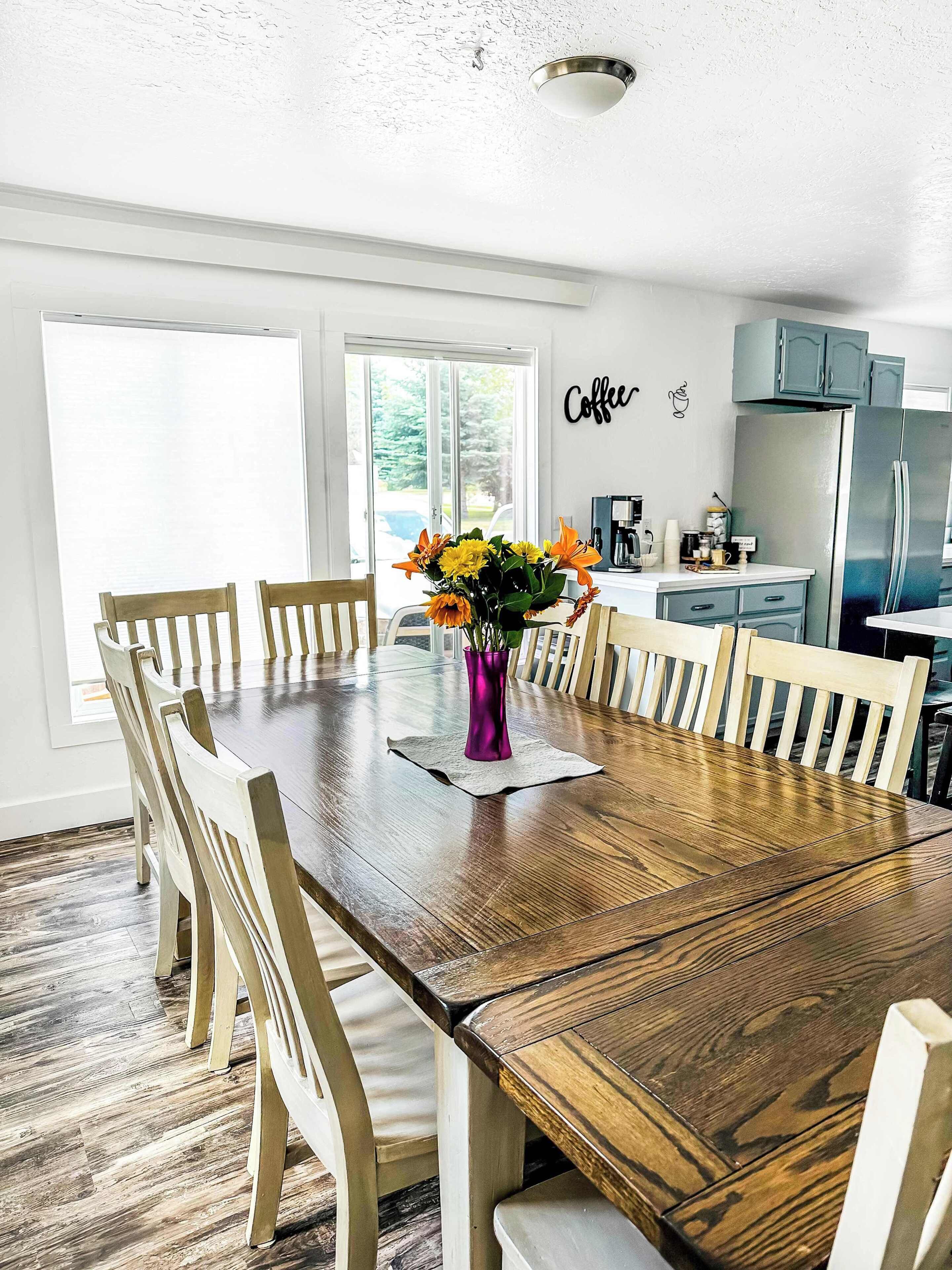 A dining area with a wooden table, white chairs, a purple vase with flowers, and a kitchen in the background.