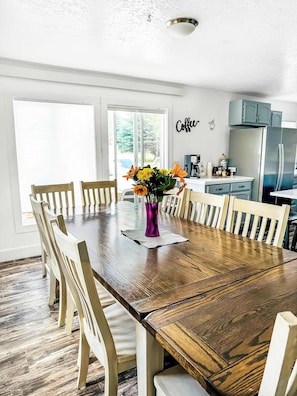 A dining area with a wooden table, white chairs, a purple vase with flowers, and a kitchen in the background.
