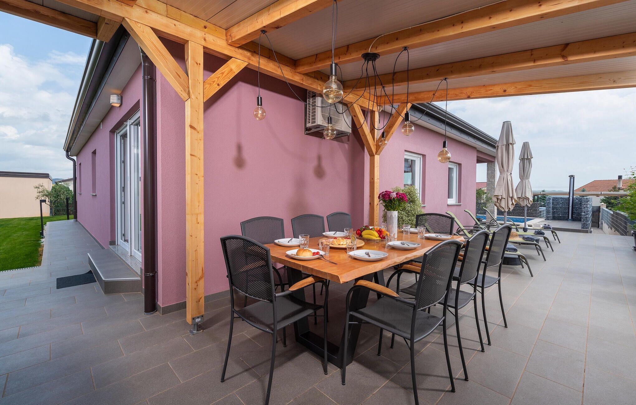 An outdoor dining area with a wooden table and chairs, a pink wall, and a view of a swimming pool.