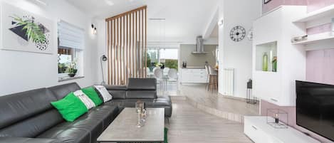 A modern living room with a leather sofa, a coffee table, and a TV. There is a kitchen in the background with a dining area.