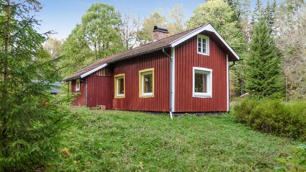 Ein rotes Holzhaus mit zwei Fenstern und einem Schornstein, umgeben von viel Grün.