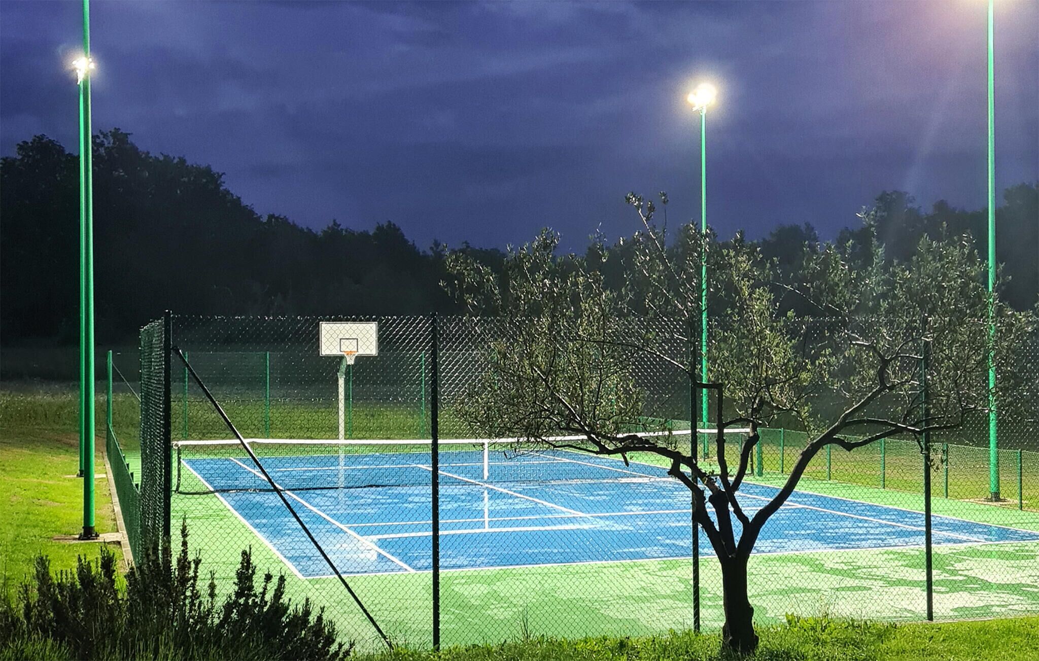 An outdoor tennis court at night with floodlights, surrounded by a chain-link fence and trees.