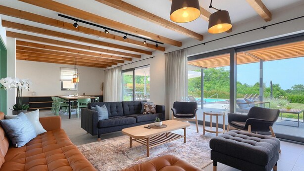 A spacious living area with a large sofa, armchairs, and a coffee table. There is a dining area with chairs and a table, and a view of a swimming pool through large windows. - Amazing home in Vizinada (Vizinada)