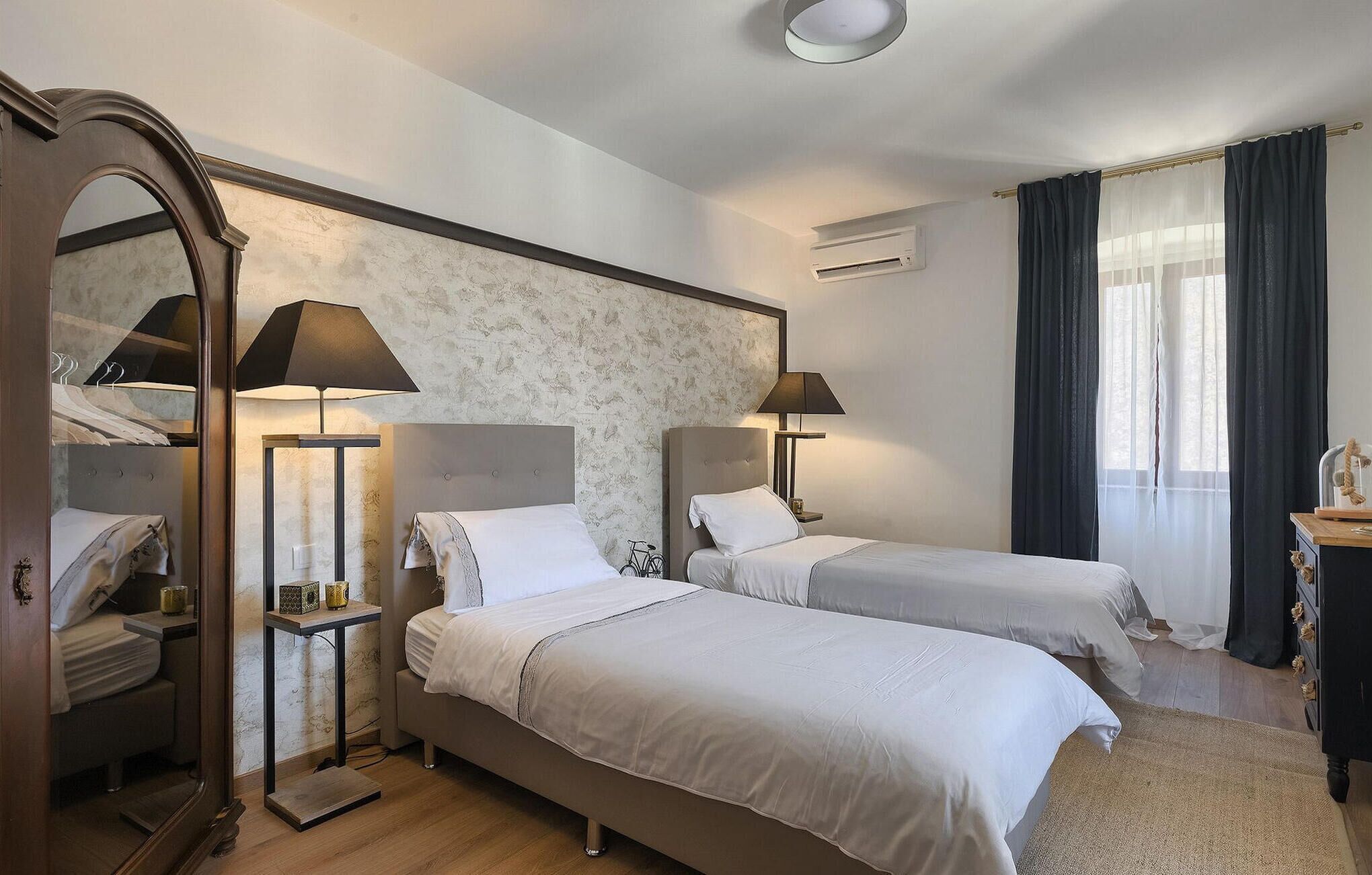 A hotel room with two beds, a mirror, a wardrobe, a small table, and a window with curtains.