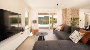 A modern living room with a large flat-screen TV, a grey sofa, and a small round table. There is a balcony with a pool and a view of greenery. - Amazing home in Labin - Rabac (Labin - Rabac)