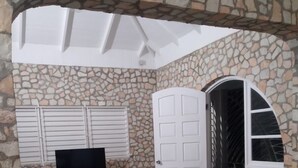 Una habitación con paredes de piedra, un sofá, un sillón de peluche rojo, un televisor sobre un mueble de madera y una puerta blanca que da a otra habitación.