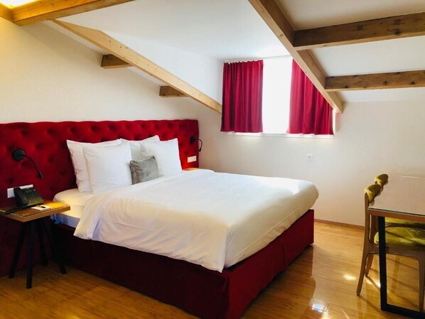 A bedroom with a red upholstered headboard, a wooden floor, a glass table with a chair, and a window with red curtains. - Chasse Residency Hotel (Amsterdam)