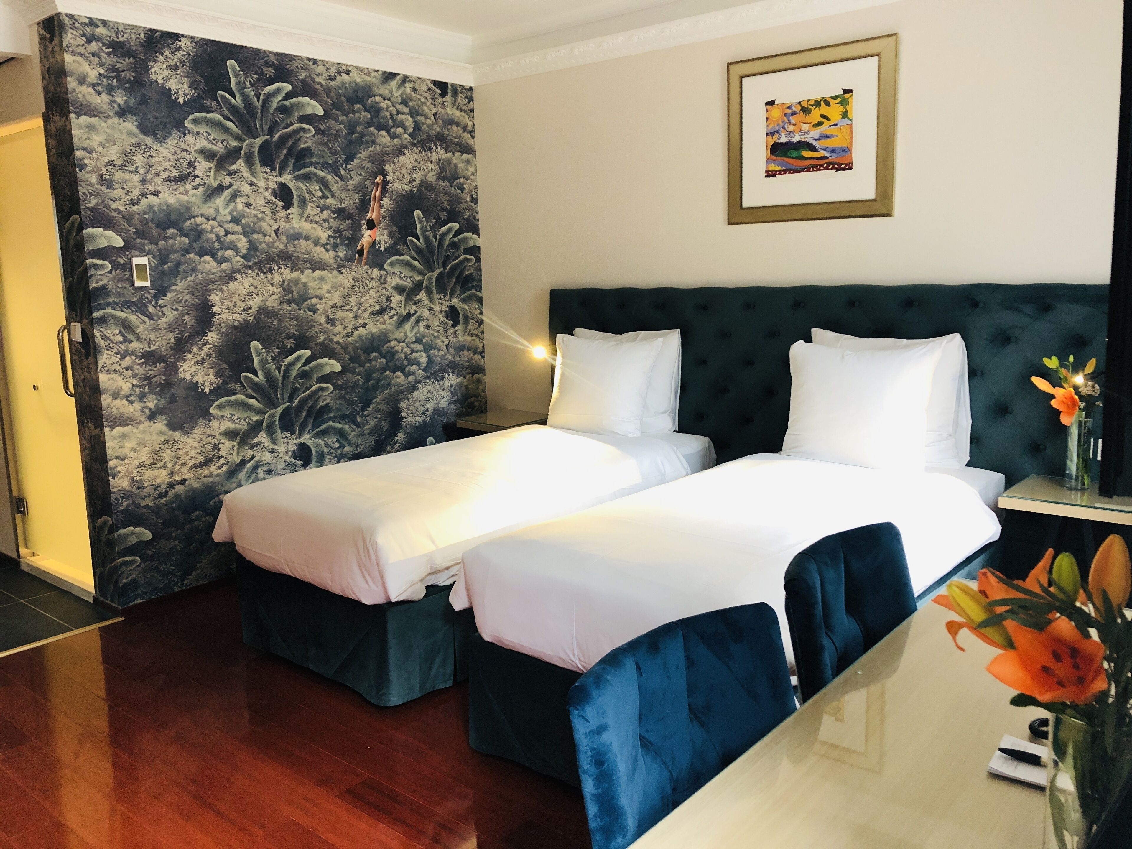 A hotel room with two beds, a dark green headboard, a mural on the wall, and a framed picture above the bed.