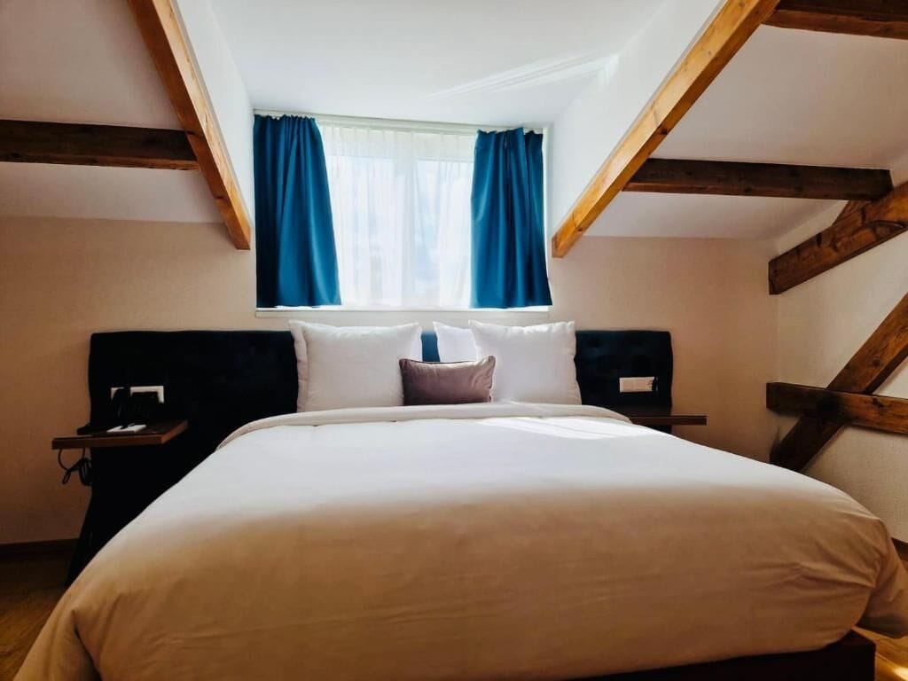 A bedroom with a large bed, wooden beams, a window with blue curtains, and a nightstand with a lamp.