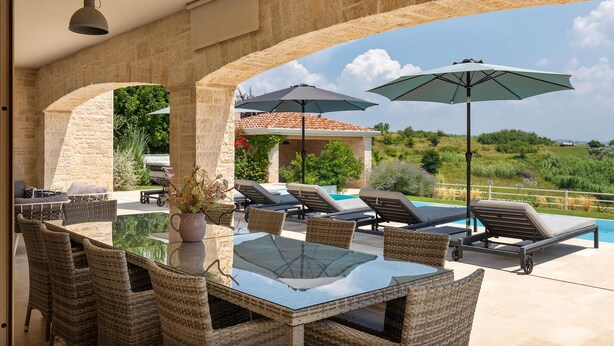 An outdoor patio with a glass-top table surrounded by wicker chairs, a pool with lounge chairs, and umbrellas. - Nice home in Porec with sauna (Porec)