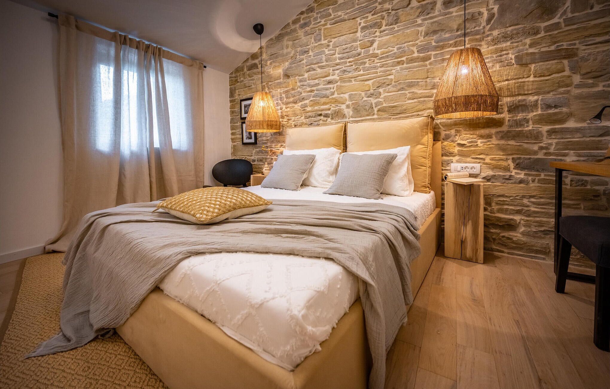Une chambre avec un mur en pierre, un lit avec une tête de lit beige, deux lampes de chevet, une table de chevet en bois et une chaise.