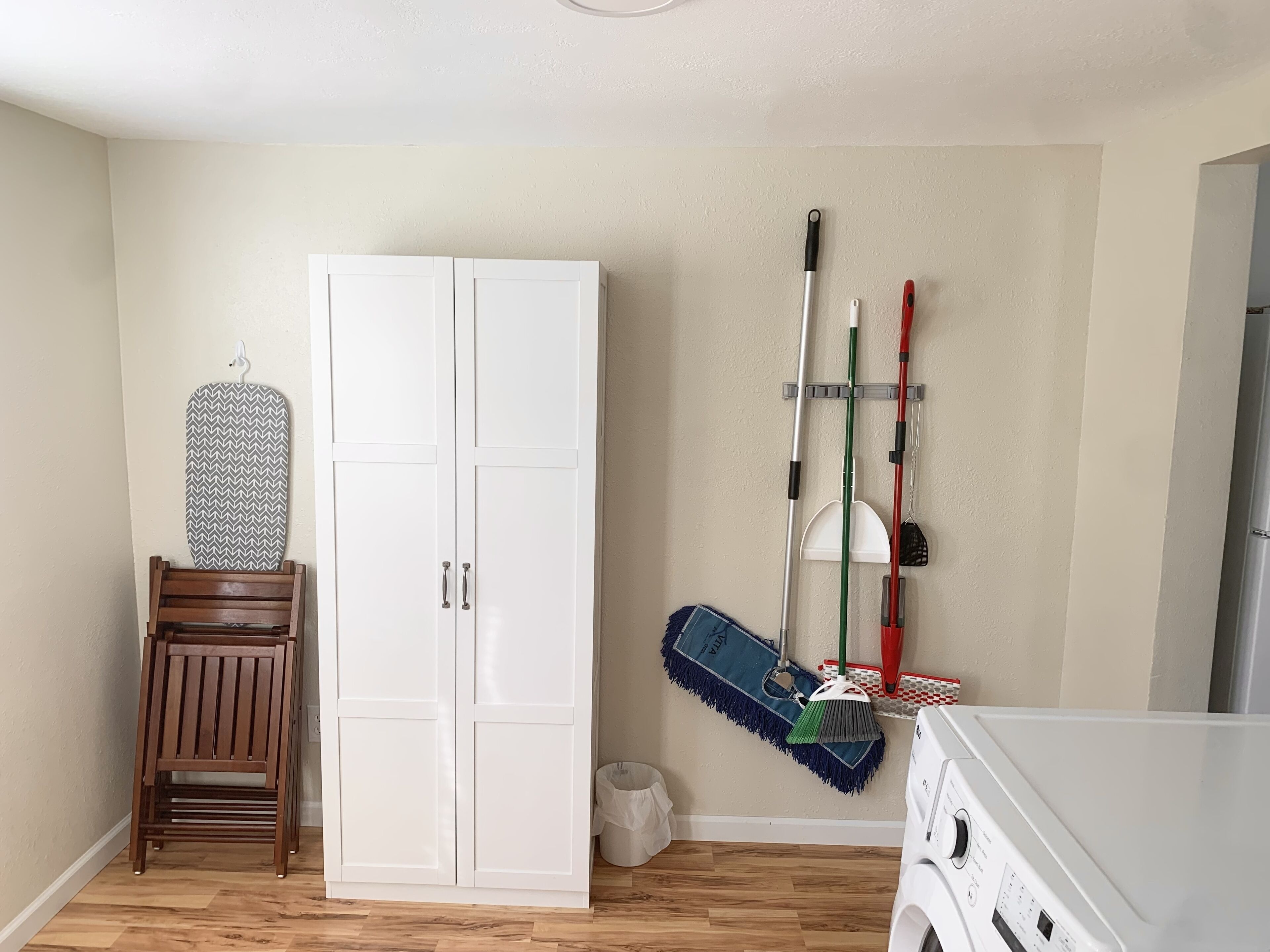 A laundry room with a white washing machine, a wooden chair, a white wardrobe, and a mop rack with various cleaning tools.
