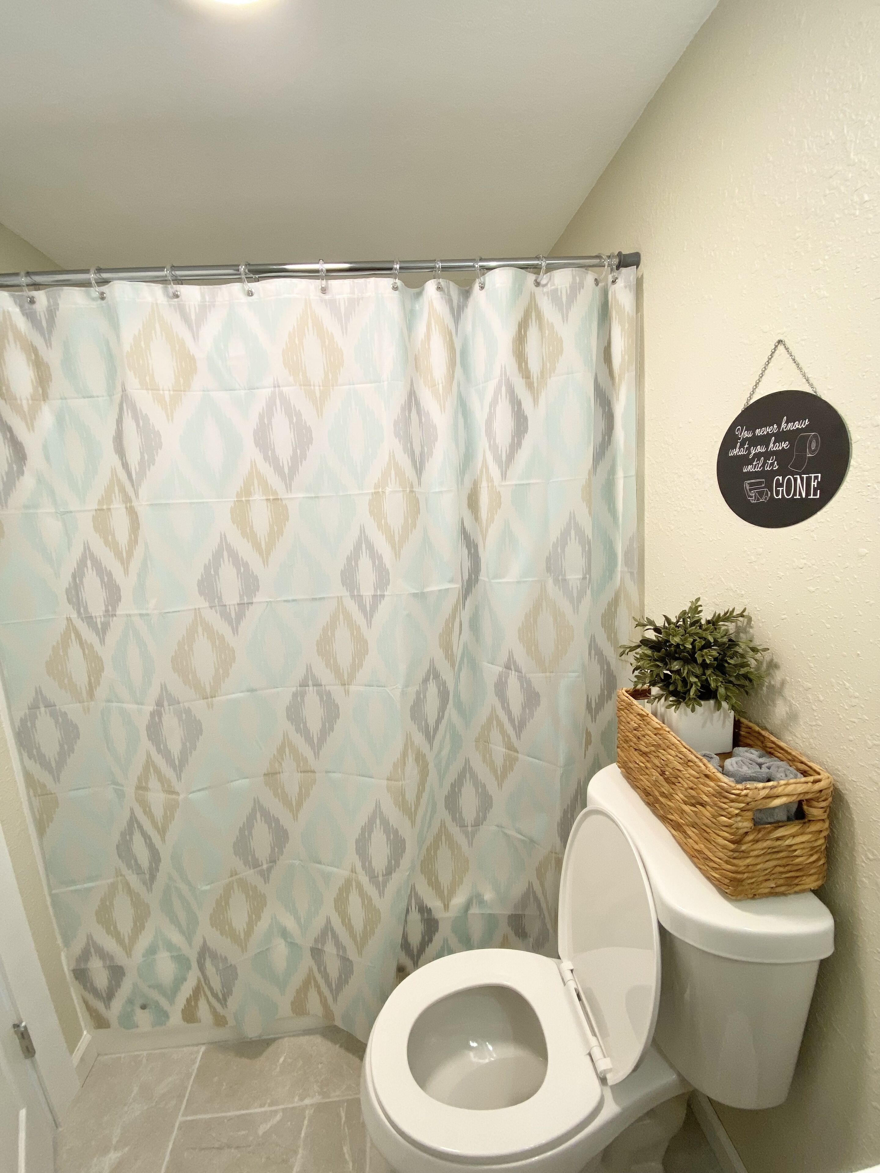 A bathroom with a patterned shower curtain, a toilet, a wicker basket with a plant, and a motivational quote on the wall.