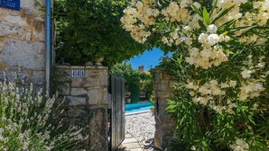 Puerta de piedra que da a una zona de piscina, rodeada de exuberante vegetación y flores blancas.