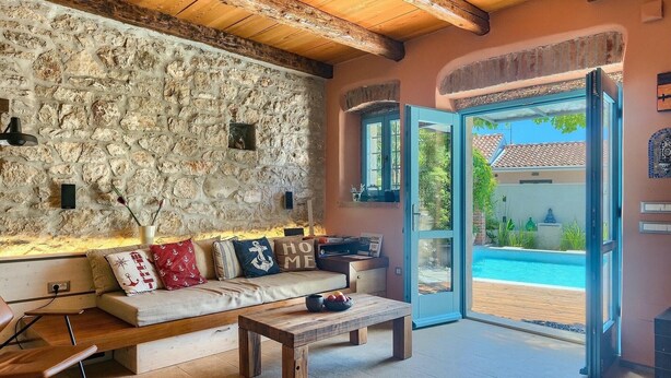 Un salón acogedor con pared de piedra, un sofá con cojines, una mesa de madera y vistas a una piscina a través de una puerta abierta.