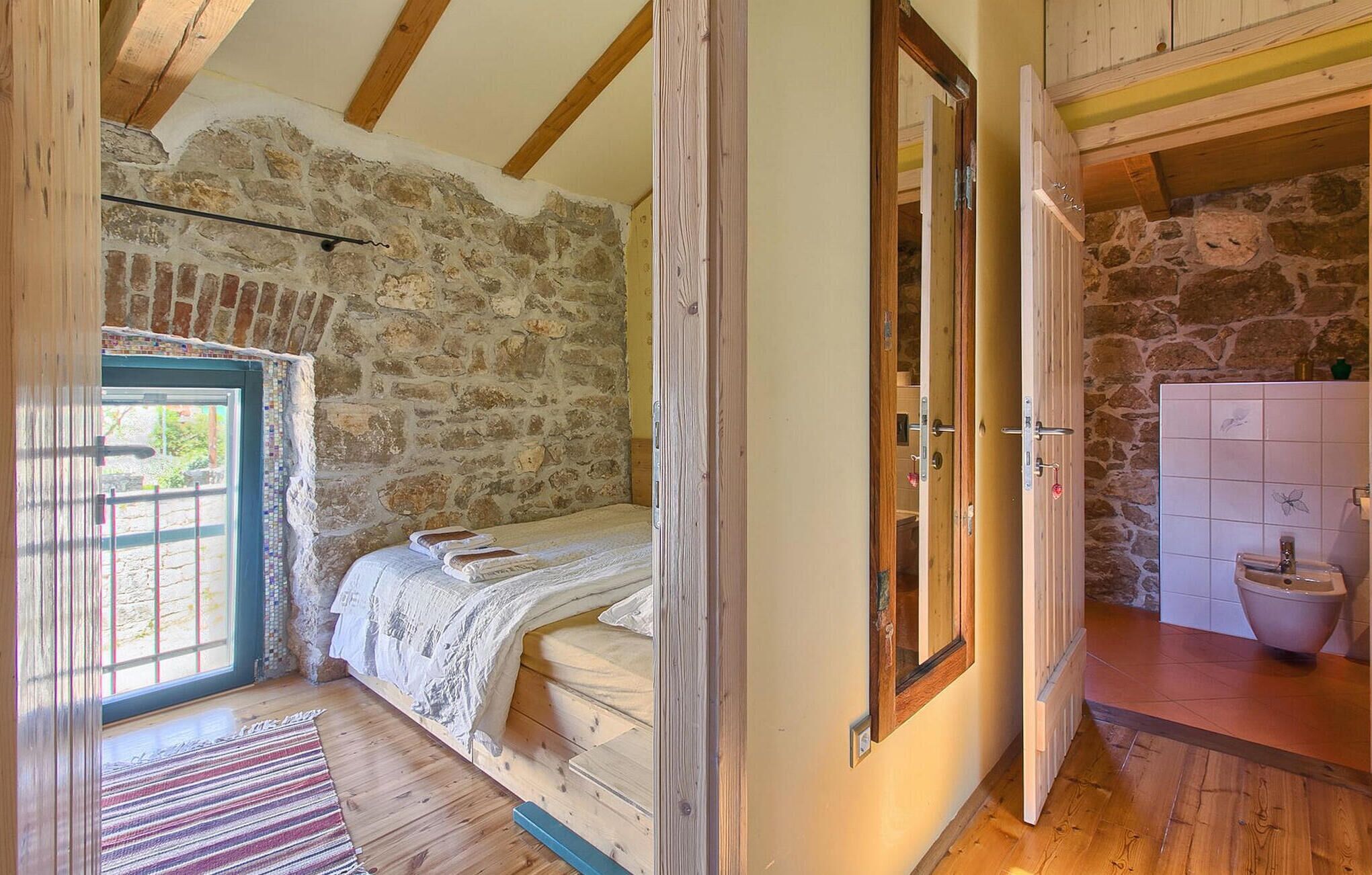 Quarto com parede de pedra, vigas de madeira, cama, porta e banheiro com pia e bacia.
