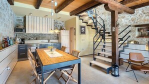 Una cocina rústica con vigas de madera, una zona de comedor con mesa de madera y una escalera que sube a otro nivel.