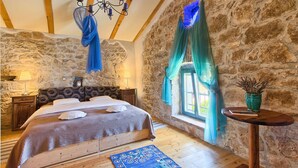 Un dormitorio con pared de piedra, una cama con colcha azul y marrón, una araña y una ventana con cortina.