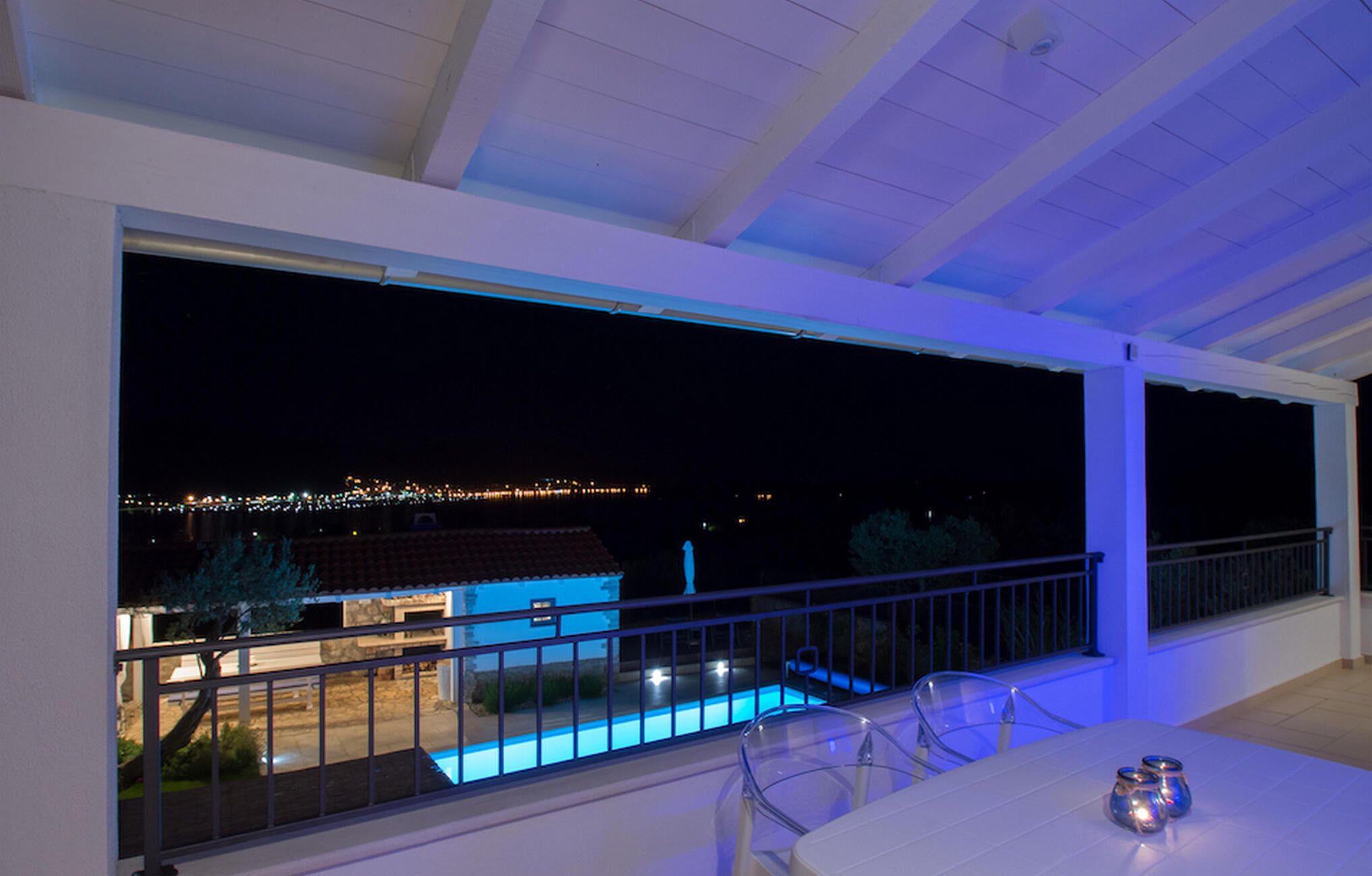 Un balcón con piscina, barandilla transparente y vista a un edificio iluminado en la distancia.