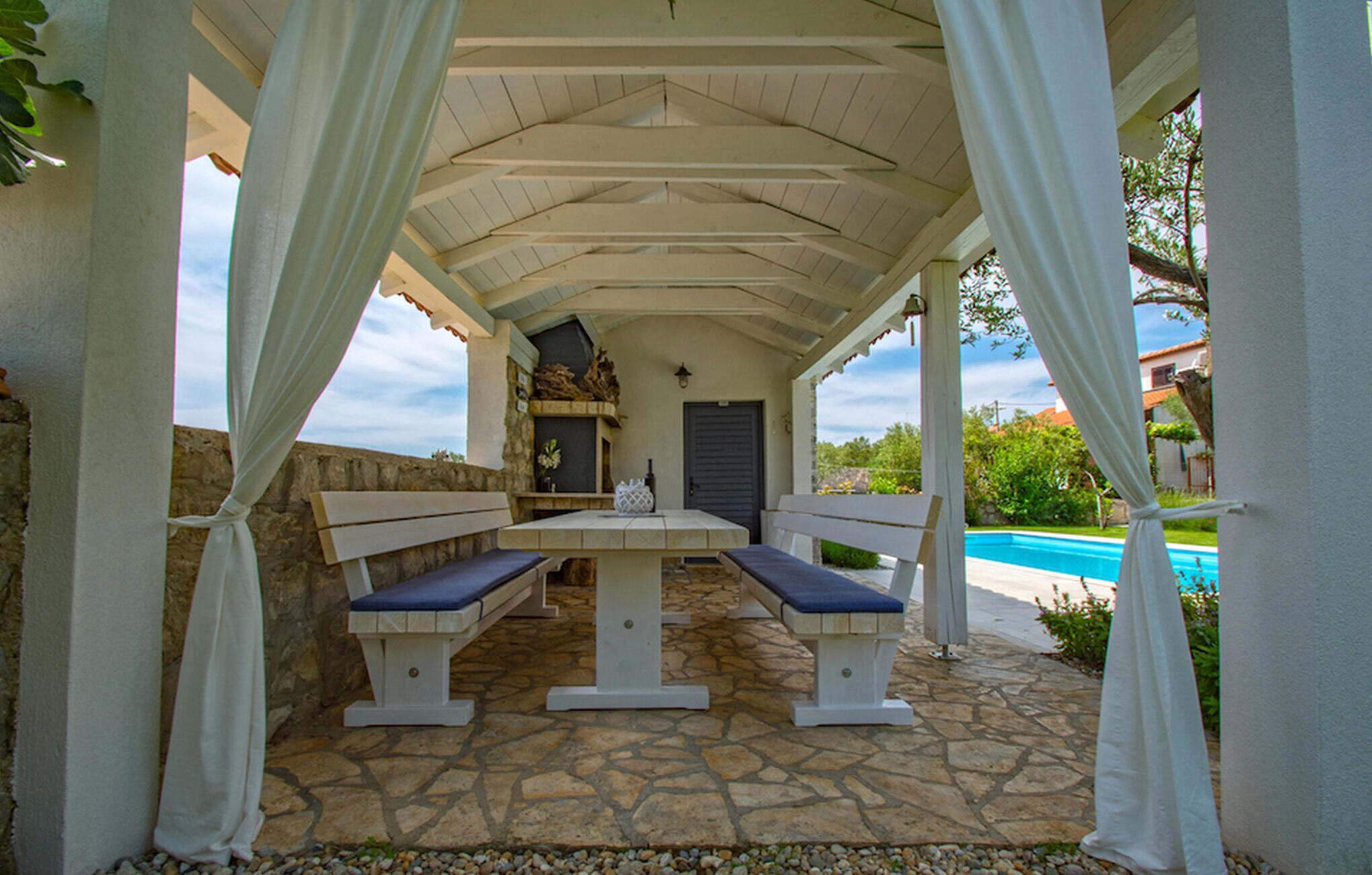Un patio exterior con suelo de piedra, chimenea de piedra y piscina al fondo.