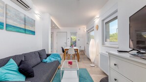 Una sala moderna con un sofá gris, una mesa de centro de vidrio y un comedor con mesa y sillas blancas. Se ve un ventanal grande a la derecha y una puerta que da a otra habitación.
