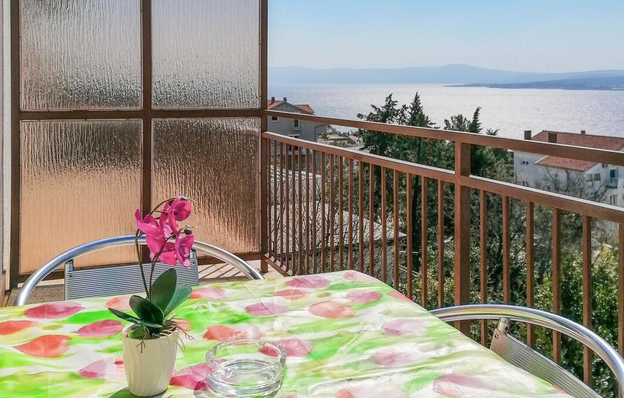 En balkong med ett bord, stolar, en krukväxt och utsikt över havet och de avlägsna bergen.
