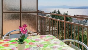 Uma varanda com mesa, cadeiras, planta em vaso e vista para o mar e montanhas ao longe.