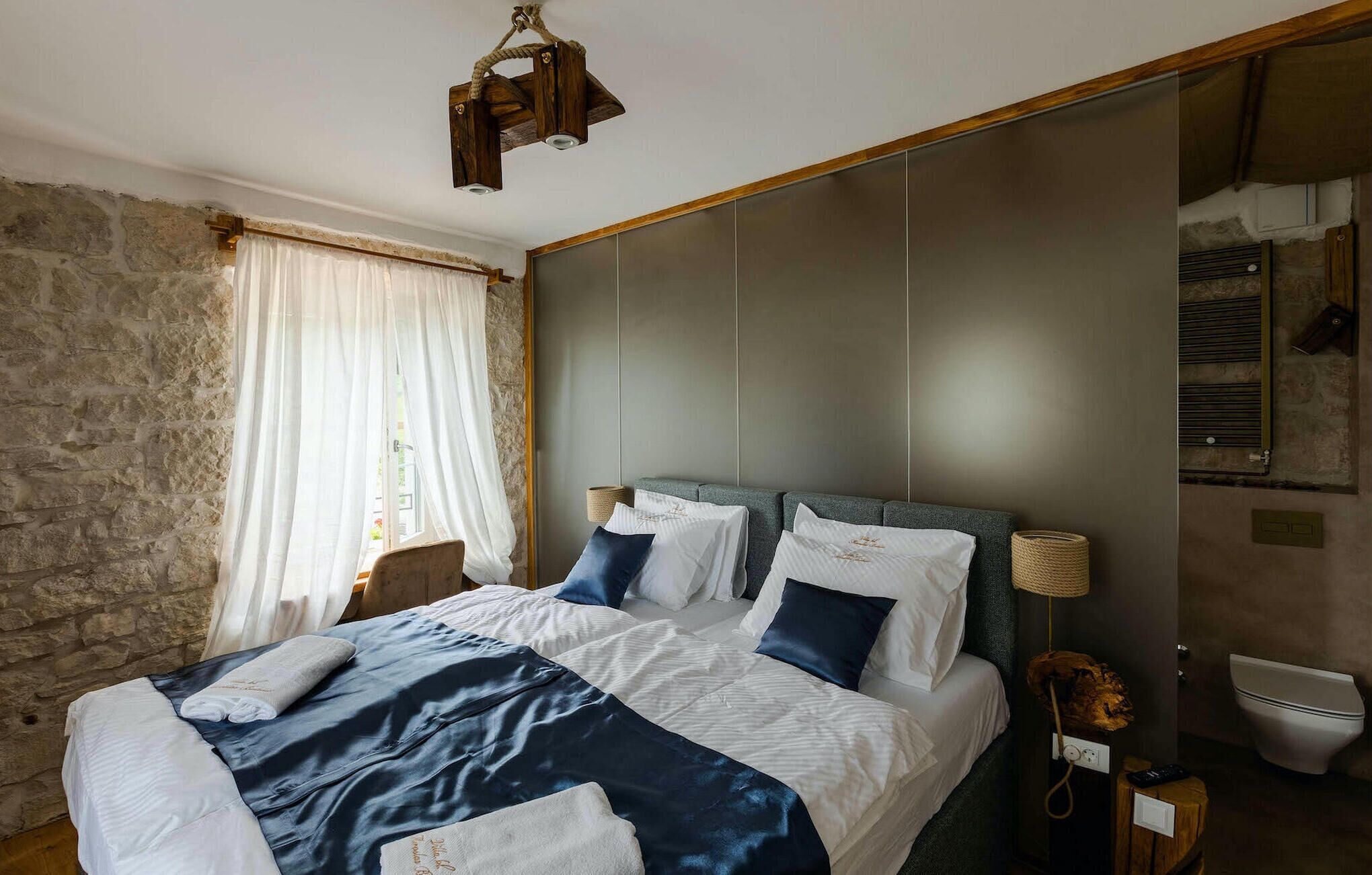 A bedroom with a stone wall, a bed with white and dark blue bedding, a wooden headboard, and a bedside lamp.