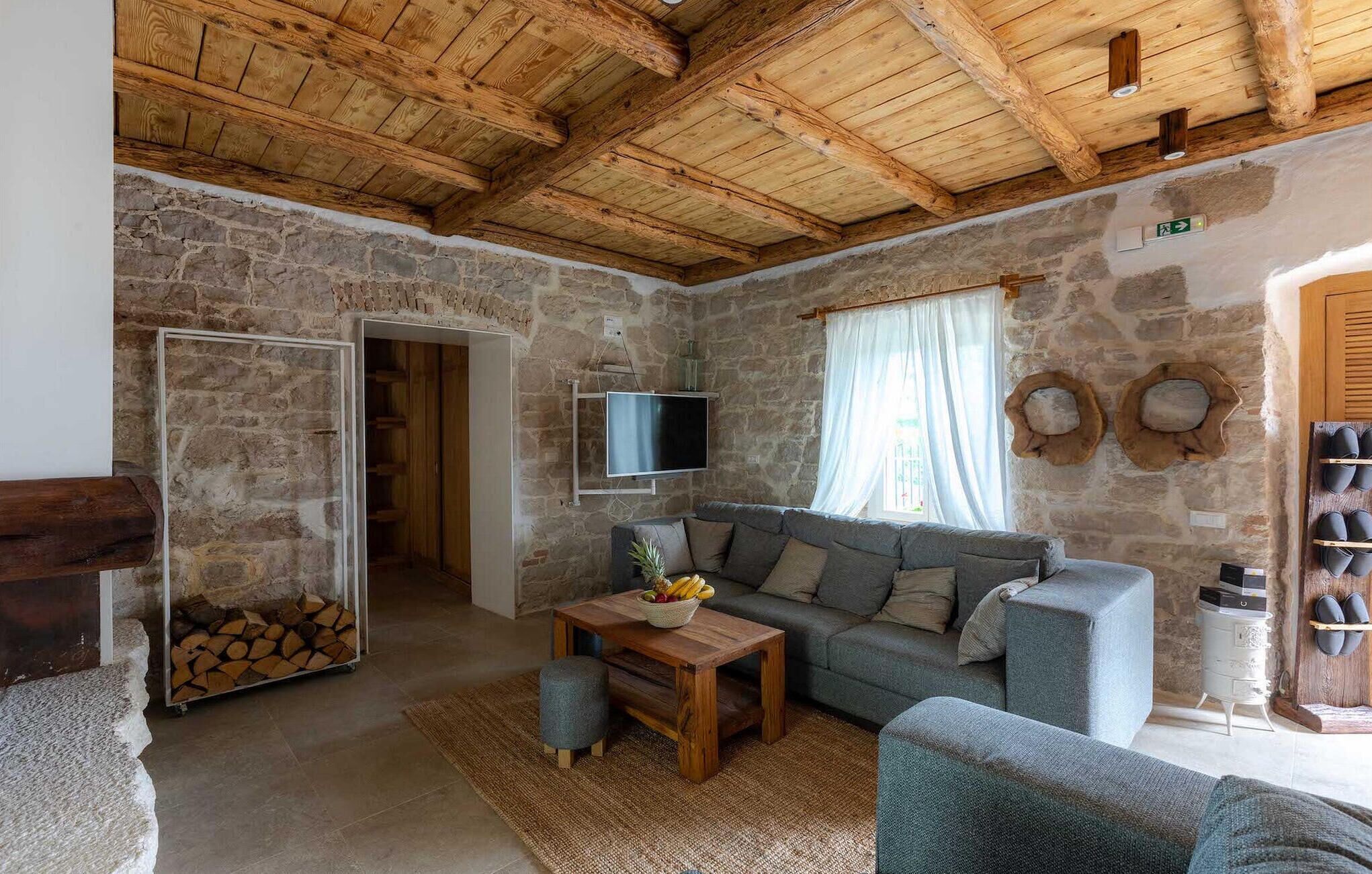 Uma sala de estar rústica com parede de pedra, vigas de madeira no teto, um sofá cinza, uma mesa de centro de madeira e uma TV fixada na parede.